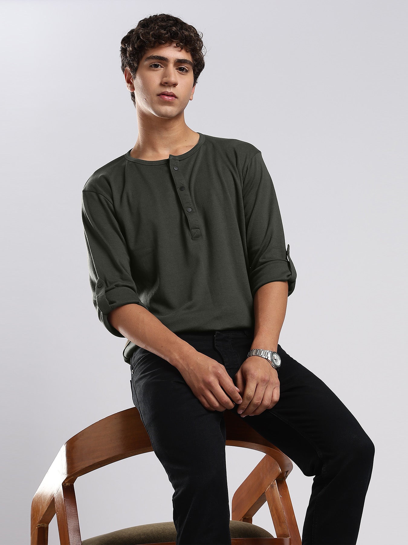 Man sitting on a wooden chair wearing a dark green shirt and black pants against a plain background