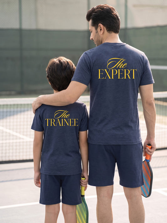 A father and son standing on a pickleball court, viewed from behind. The father wears a navy blue t-shirt with 'The EXPERT' printed in gold script and the son wears a matching navy blue t-shirt with 'The TRAINEE' printed in gold script.