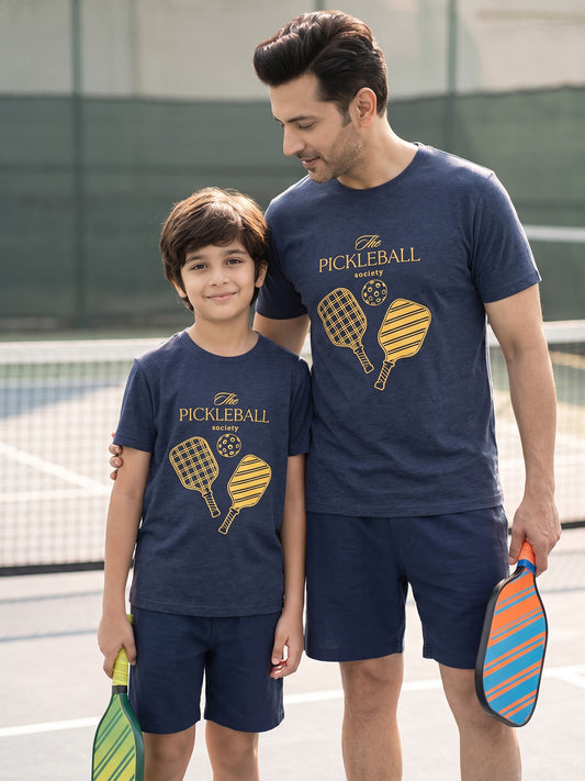 A man and a boy wearing matching navy blue pickleball t-shirts and shorts sets on an outdoor pickleball court. The navy blue shirts feature a yellow and gold 'PICKLEBALL' graphic with crossed paddles.