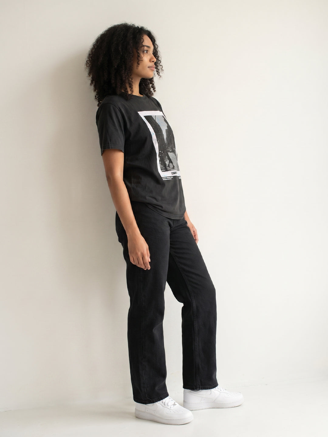 Person wearing a black t-shirt with a graphic design, black pants, and white shoes against a light background