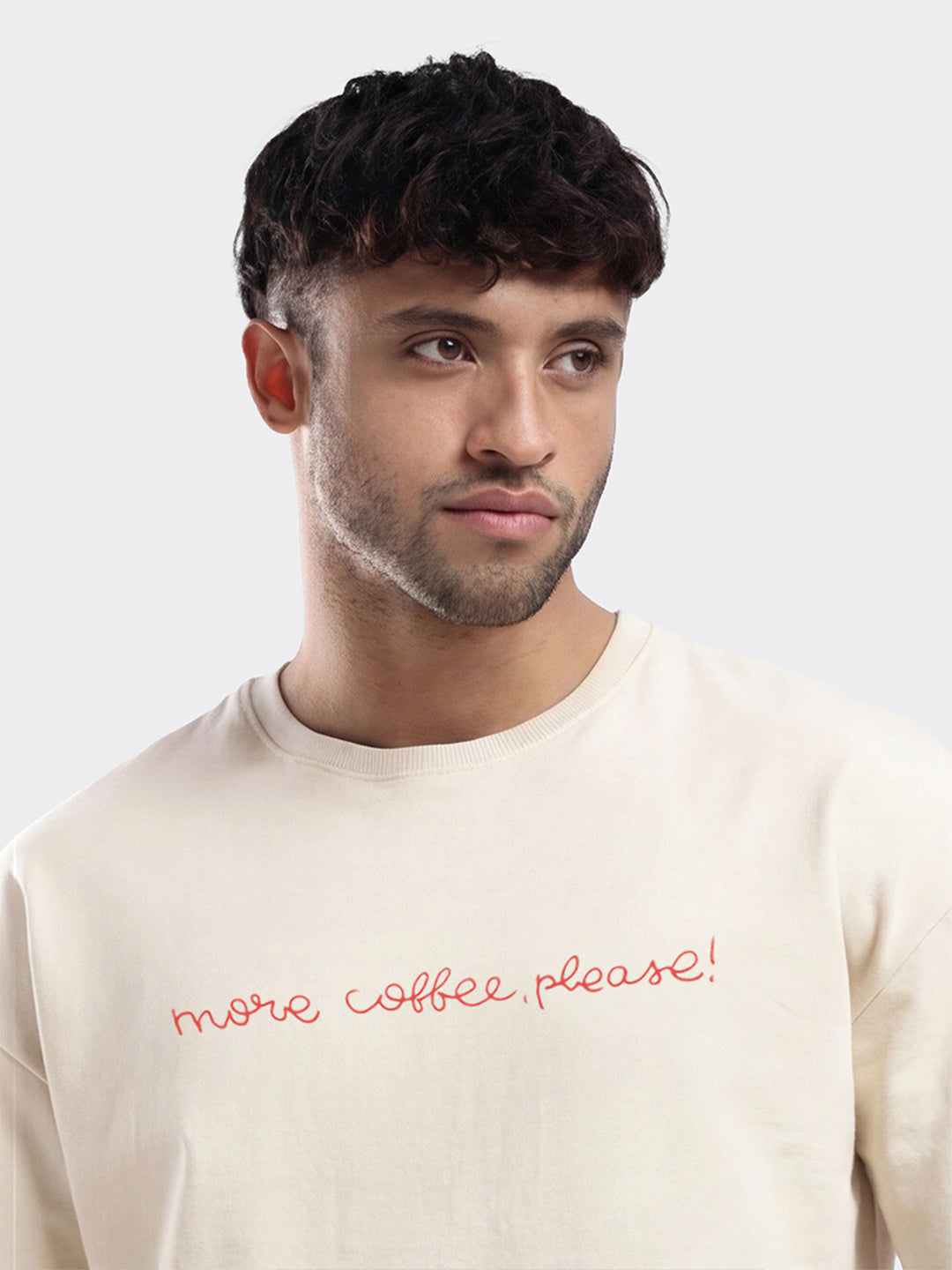Man wearing a beige sweatshirt with 'more coffee, please!' text on a plain background