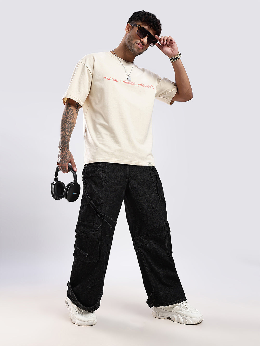 Man wearing a beige t-shirt with text, black pants, and holding headphones on a white background