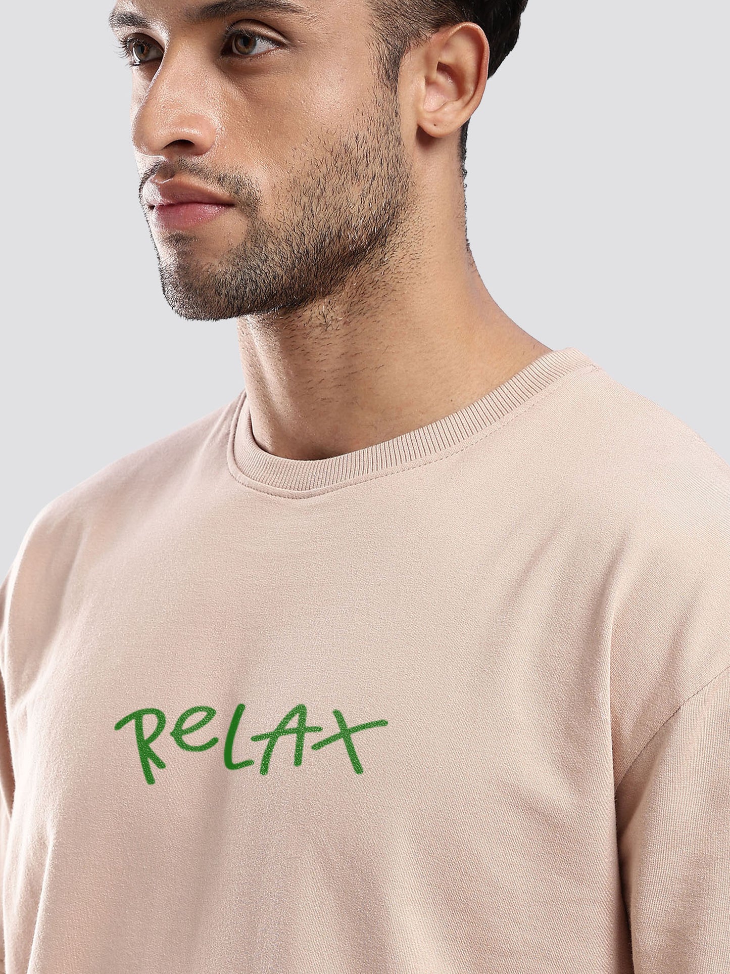 Man wearing a beige oversized t-shirt  with 'RELAX' in green text on a light gray background