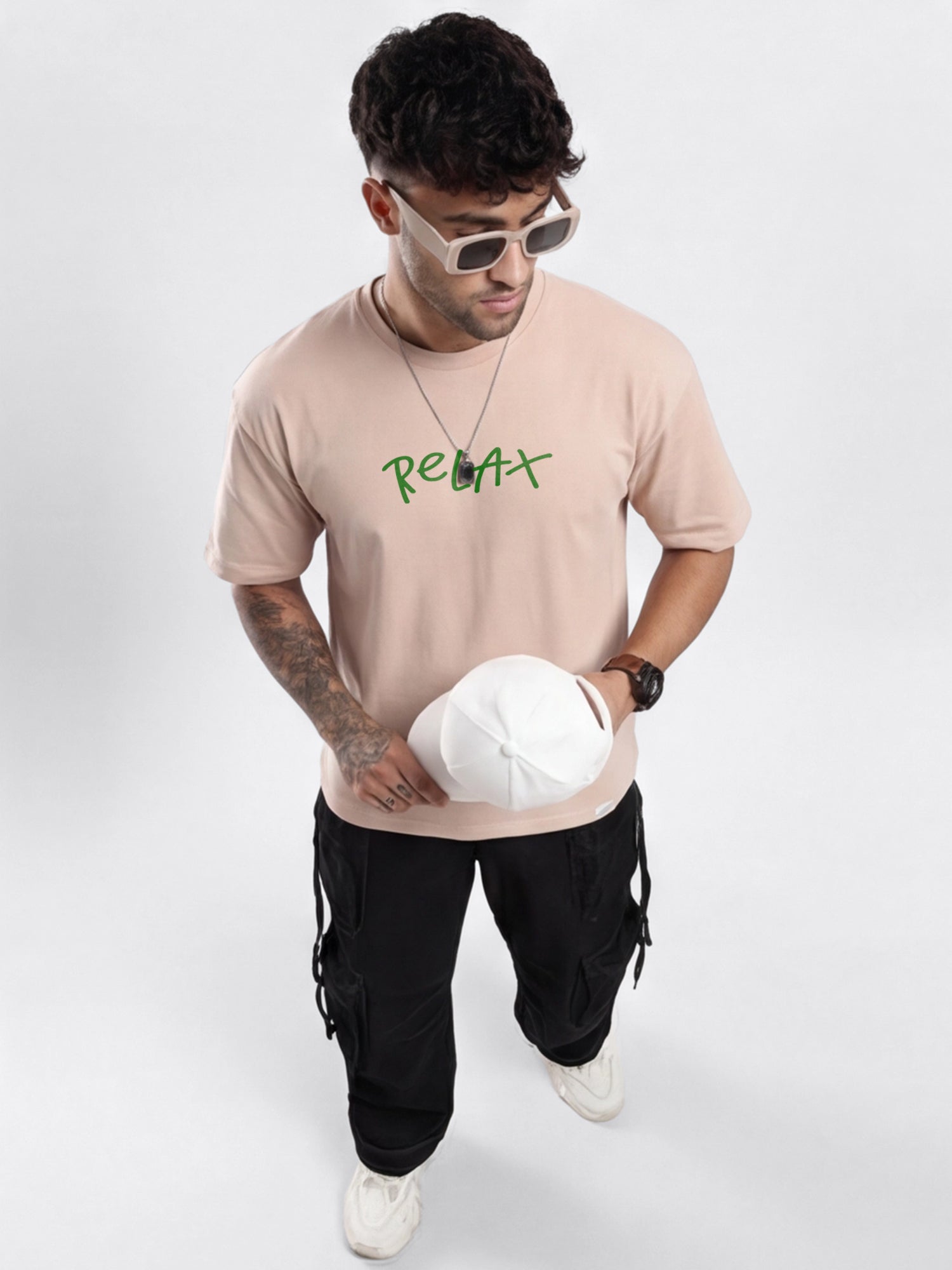 Man wearing a beige oversized t-shirt with text, black pants, and white sneakers on a white background