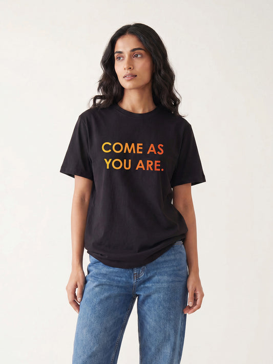 Indian female model wearing Keos Come As You Are black graphic t-shirt with yellow orange text print, styled with blue denim jeans.
