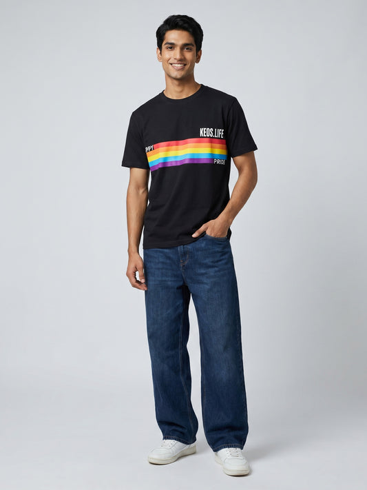 Keos Happy Pride black cotton graphic t-shirt featuring rainbow Pride stripe artwork, styled with wide-leg blue denim and white sneakers.