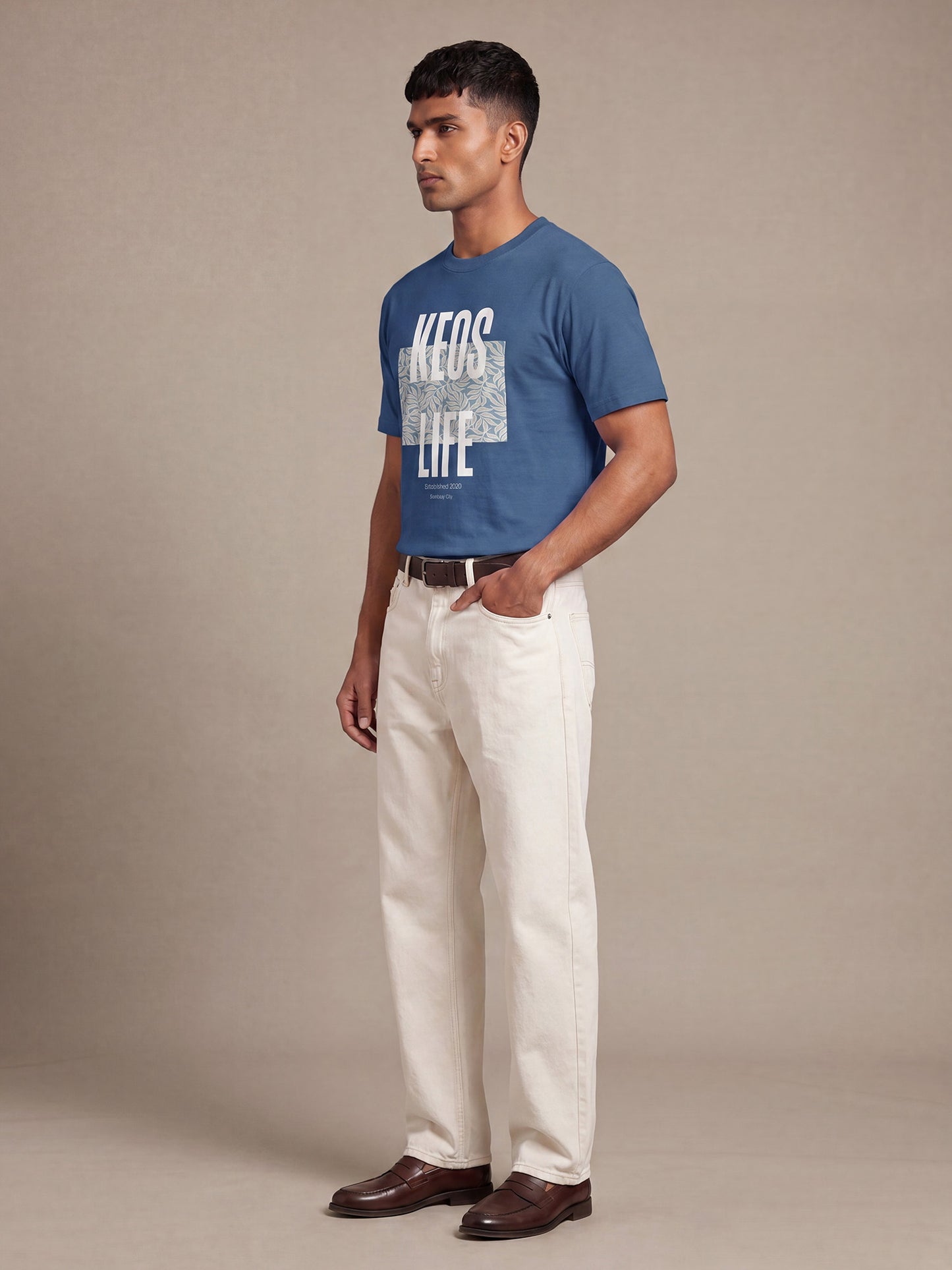 Side profile of men’s Keos Floral Block blue graphic tee with white front print, styled with cream straight-fit jeans and brown shoes.
