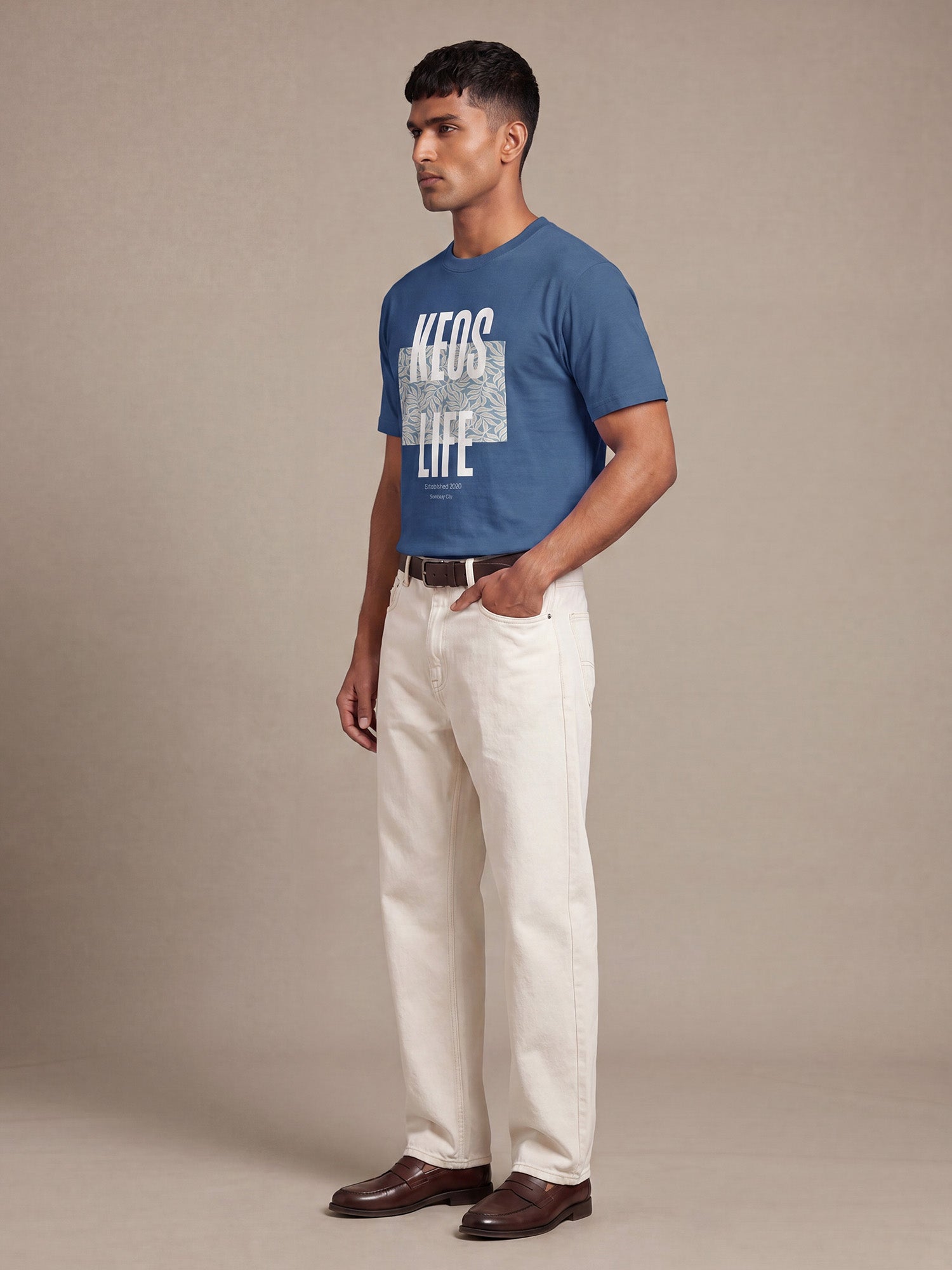 Side profile of men’s Keos Floral Block blue graphic tee with white front print, styled with cream straight-fit jeans and brown shoes.