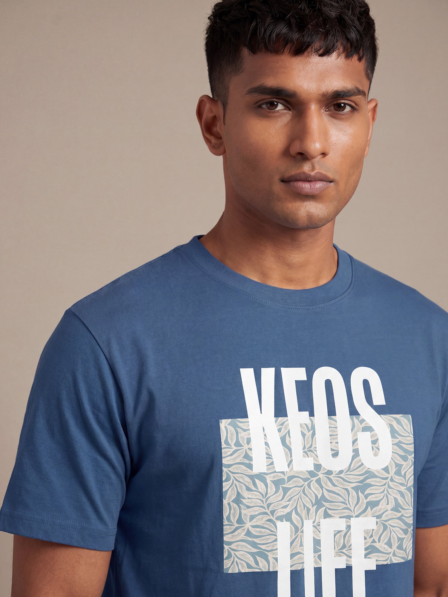 Close-up of Keos Floral Block blue cotton t-shirt showing white KEOS LIFE print, floral block artwork, crew neck, and soft fabric texture.