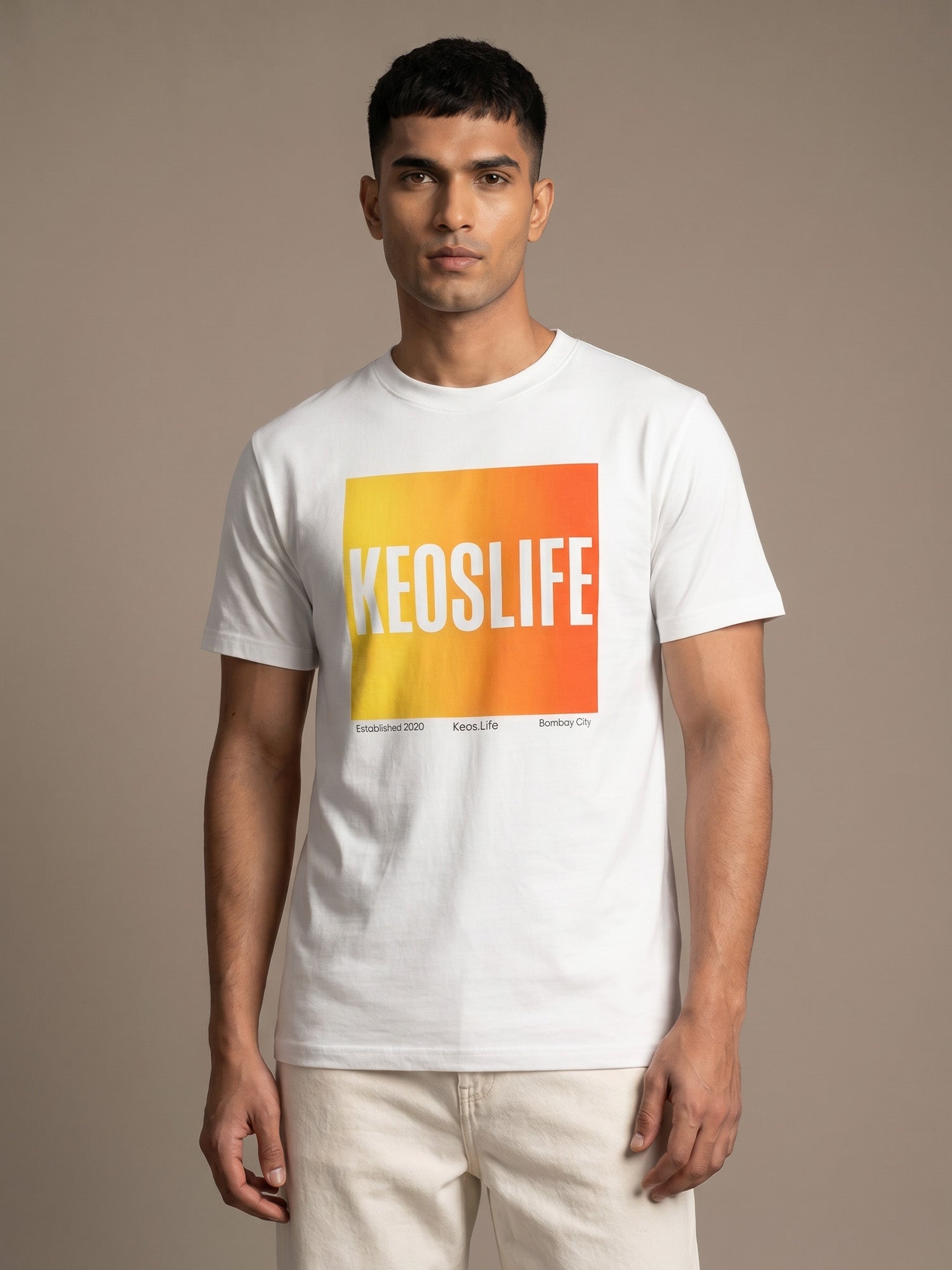 Indian male model wearing Keos Pride Block white graphic t-shirt with yellow orange KEOSLIFE print, styled with off-white trousers.