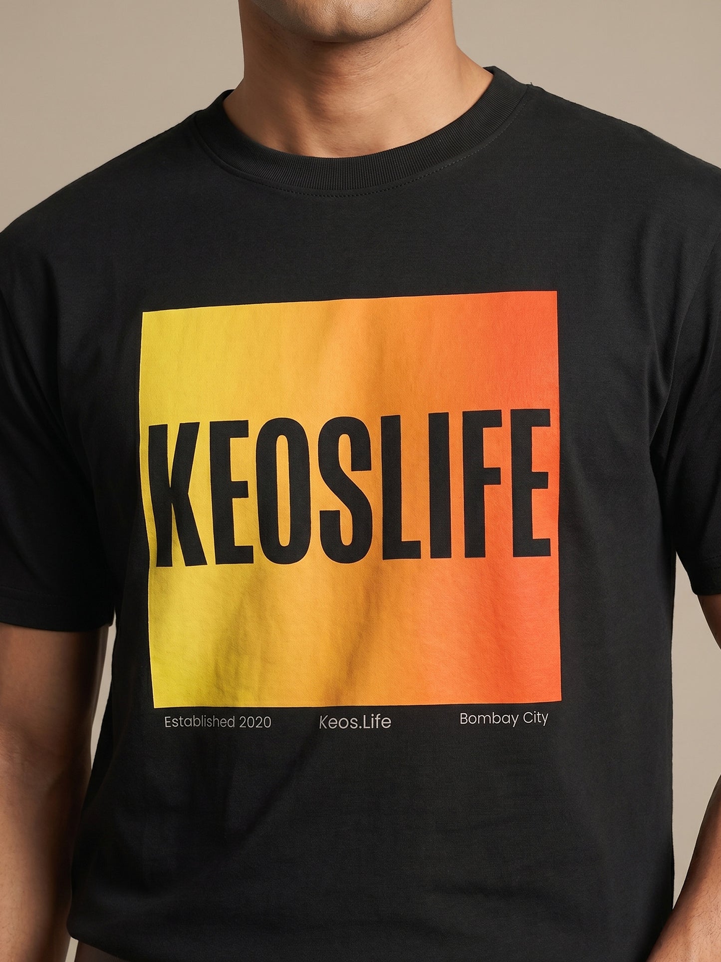 Close-up of Keos Pride Block black cotton t-shirt showing yellow orange KEOSLIFE graphic print, ribbed crew neck, and fabric texture.