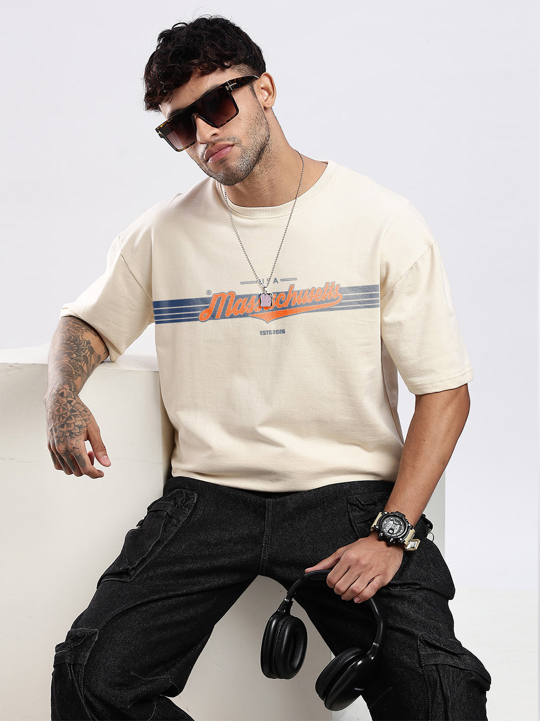 Man wearing a beige t-shirt with chest print, sunglasses, and black pants sitting on a white box.