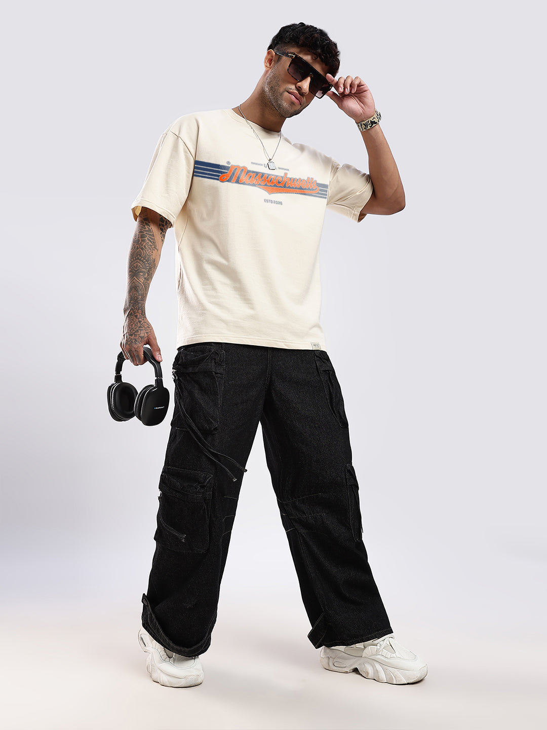 Person wearing a beige t-shirt with text, black pants, and holding headphones on a white background
