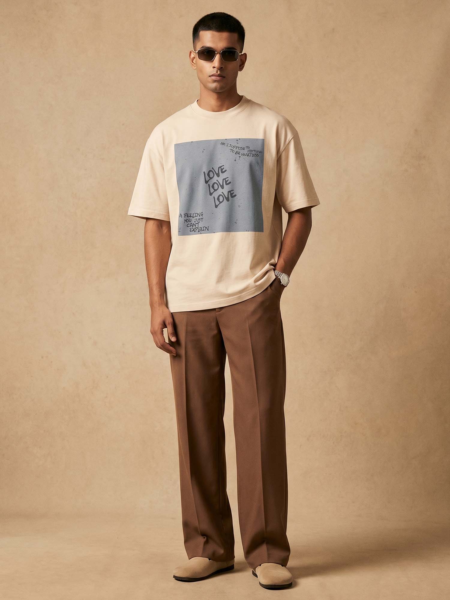 Man wearing a beige t-shirt with graphic design and brown pants against a beige background