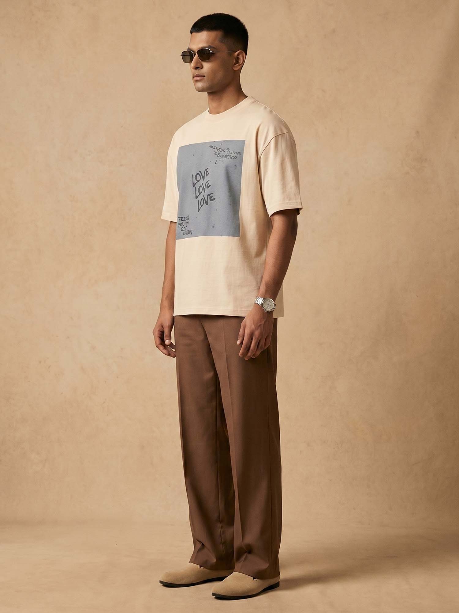 Man wearing a beige t-shirt with a graphic design and brown pants against a beige background