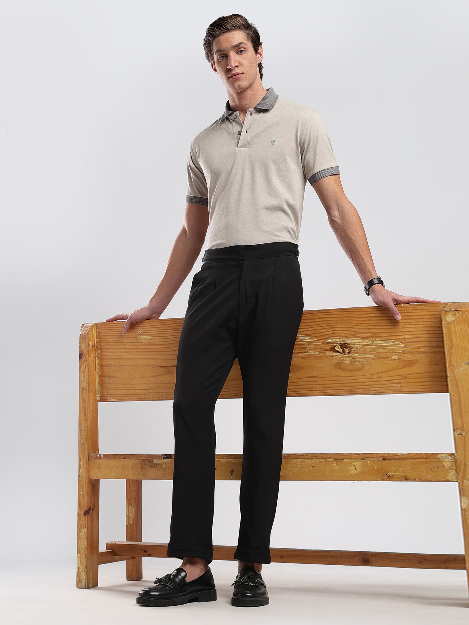 Man wearing a beige polo shirt and black pants standing against a white background with a wooden bench.