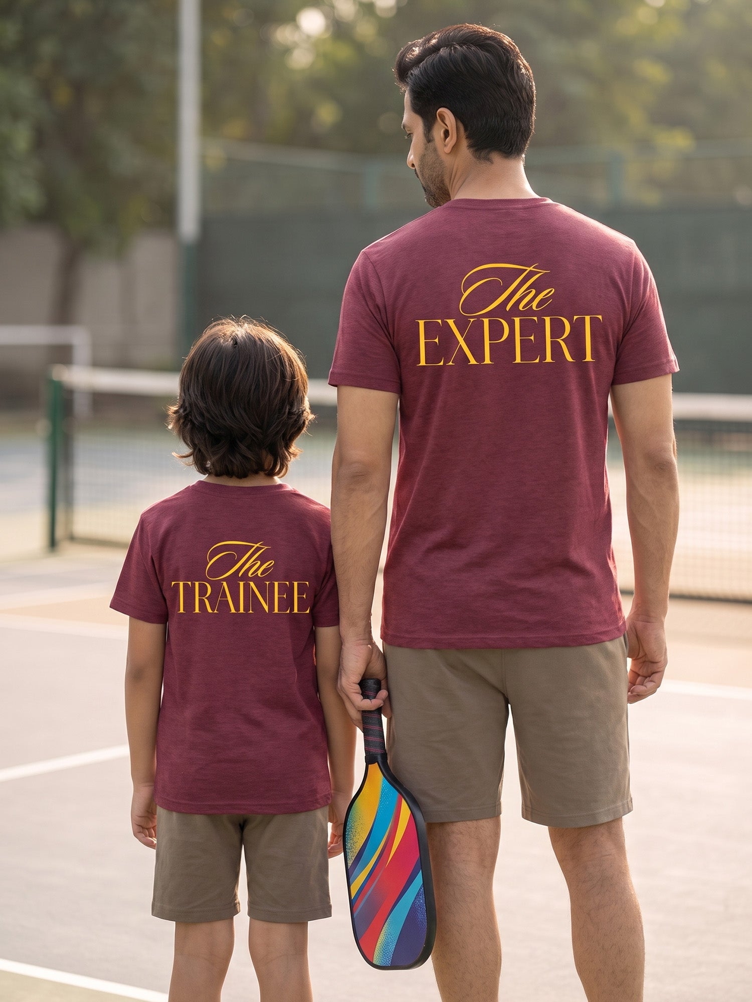 A man and a child standing on a pickleball court, both wearing matching maroon t-shirts with gold text on the back. The man's shirt reads 'The EXPERT' and the child's shirt reads 'The TRAINEE'.