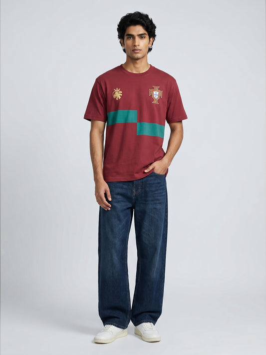 Man wearing a maroon t-shirt with a green stripe and logo of Portugal football  , paired with blue jeans on a white background