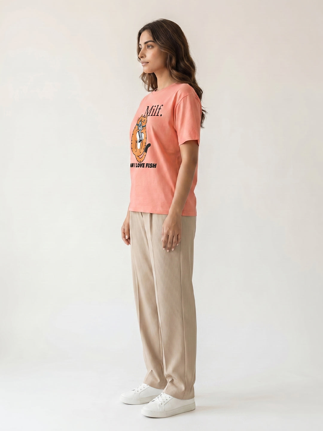 Woman wearing a peach t-shirt with text and graphics, beige pants, and white sneakers on a white background